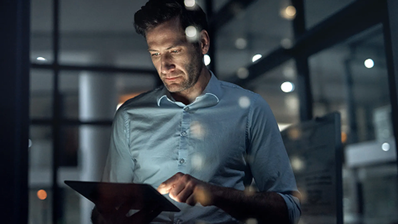 A man in a blue shirt uses a tablet in a dimly lit office, with reflections of lights visible on the glass behind him.