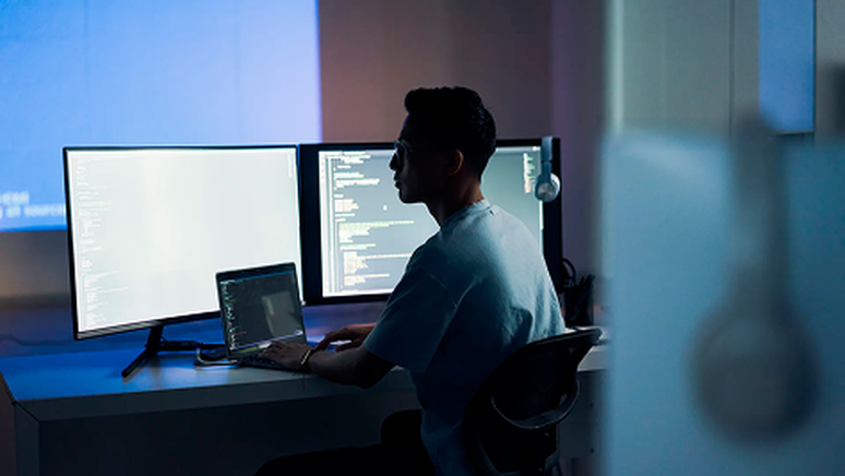 A person sits at a desk with multiple monitors displaying code, working on a laptop in a dimly lit room.