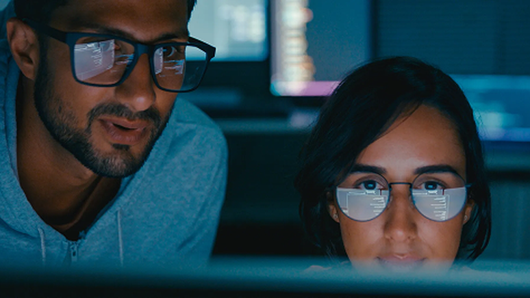 Two people wearing glasses focus on computer screens in a dimly lit room, with code reflecting on their lenses.