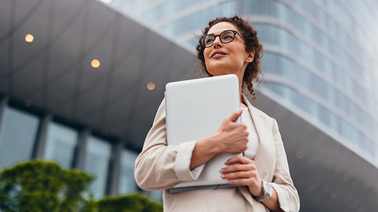A woman in a light suit and glasses holds a laptop, standing confidently in front of a modern glass building.