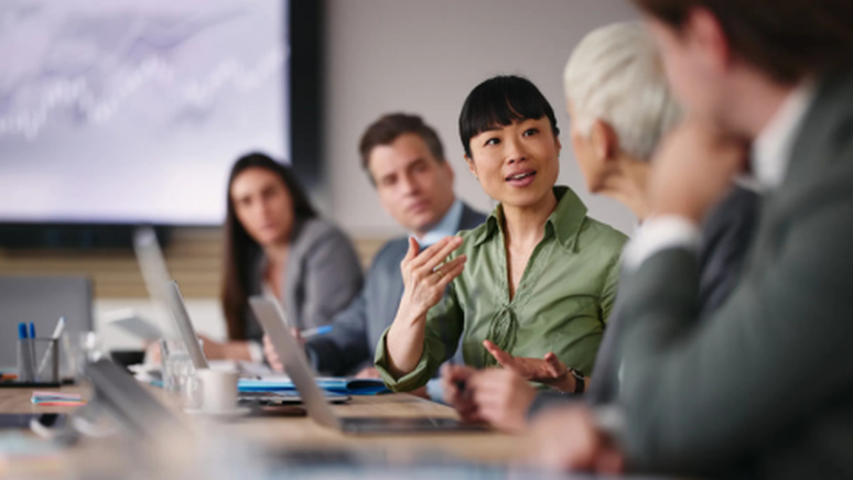 A woman speaks in a business meeting, with colleagues listening attentively around a conference table, and a screen displaying a graph in the background.