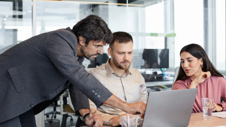 Three colleagues collaborating at a desk in a modern office, with one man pointing at a laptop screen while the others look on attentively.