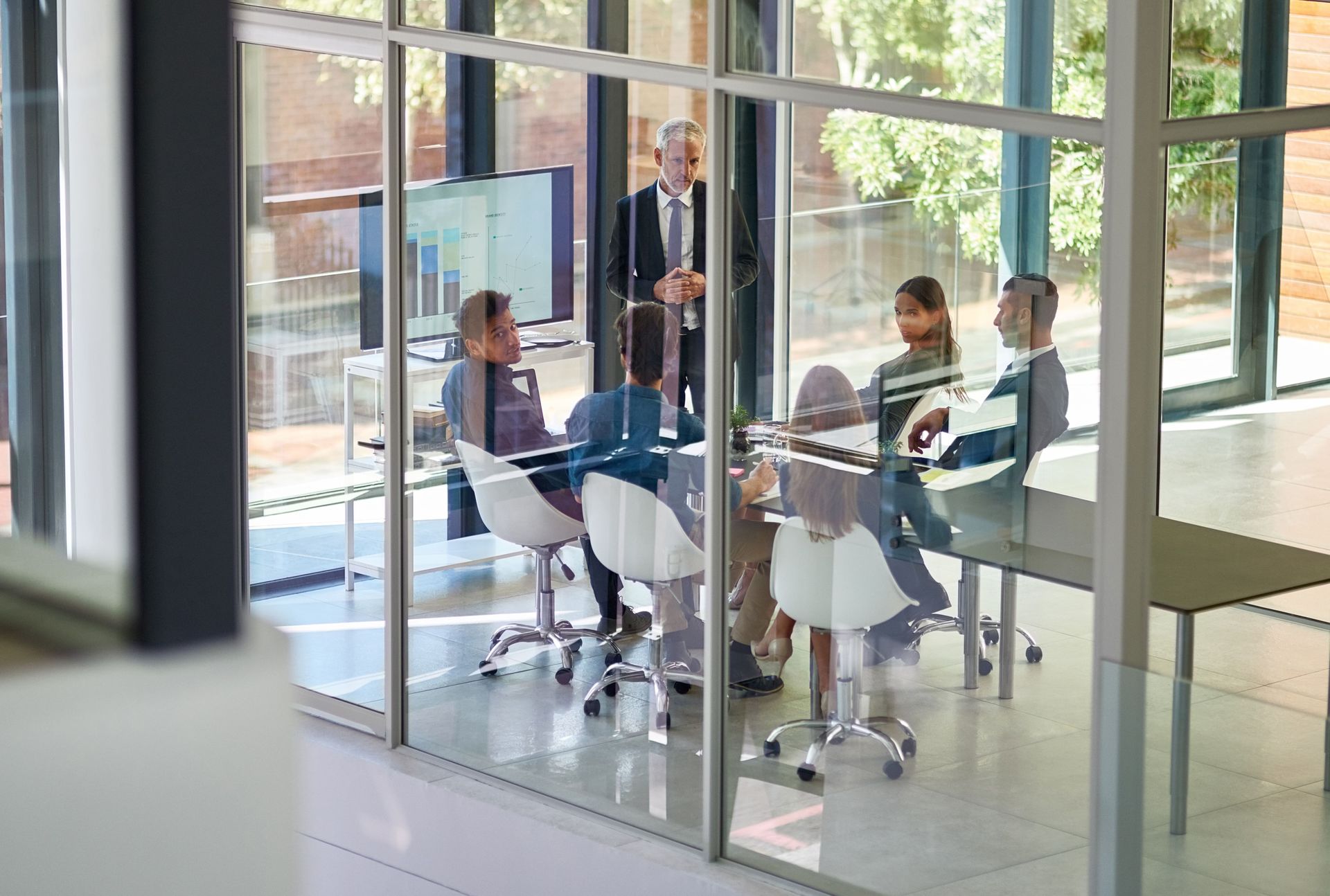 Employees in a glass-walled meeting room engaged in discussion. A presenter stands near a screen displaying data.