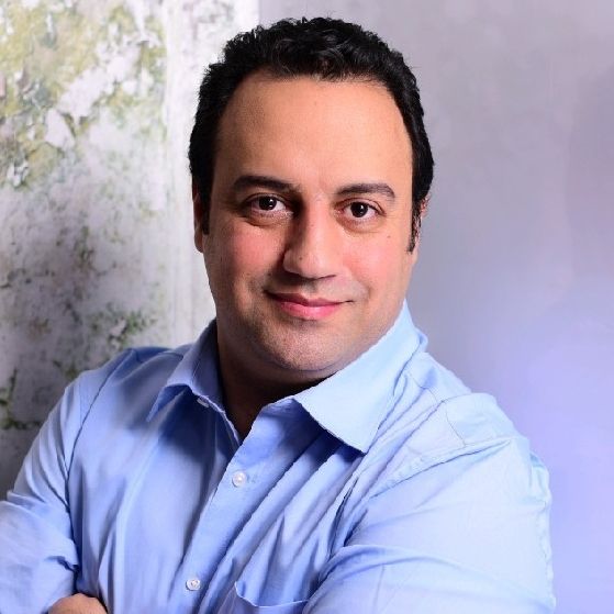 Man in a light blue shirt smiles with arms crossed, standing against a textured background.