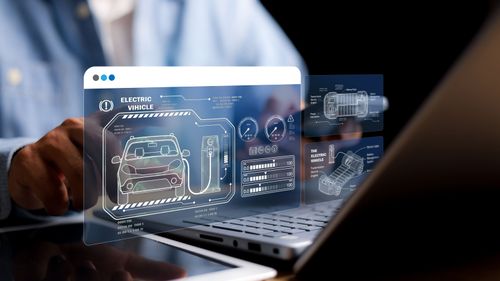 Person interacts with a holographic electric vehicle interface, displaying schematics and data over a laptop.