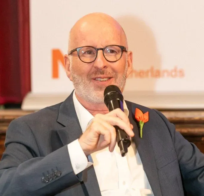 Bald man with glasses holding a microphone, wearing a suit with an orange flower pin, smiling during an event.