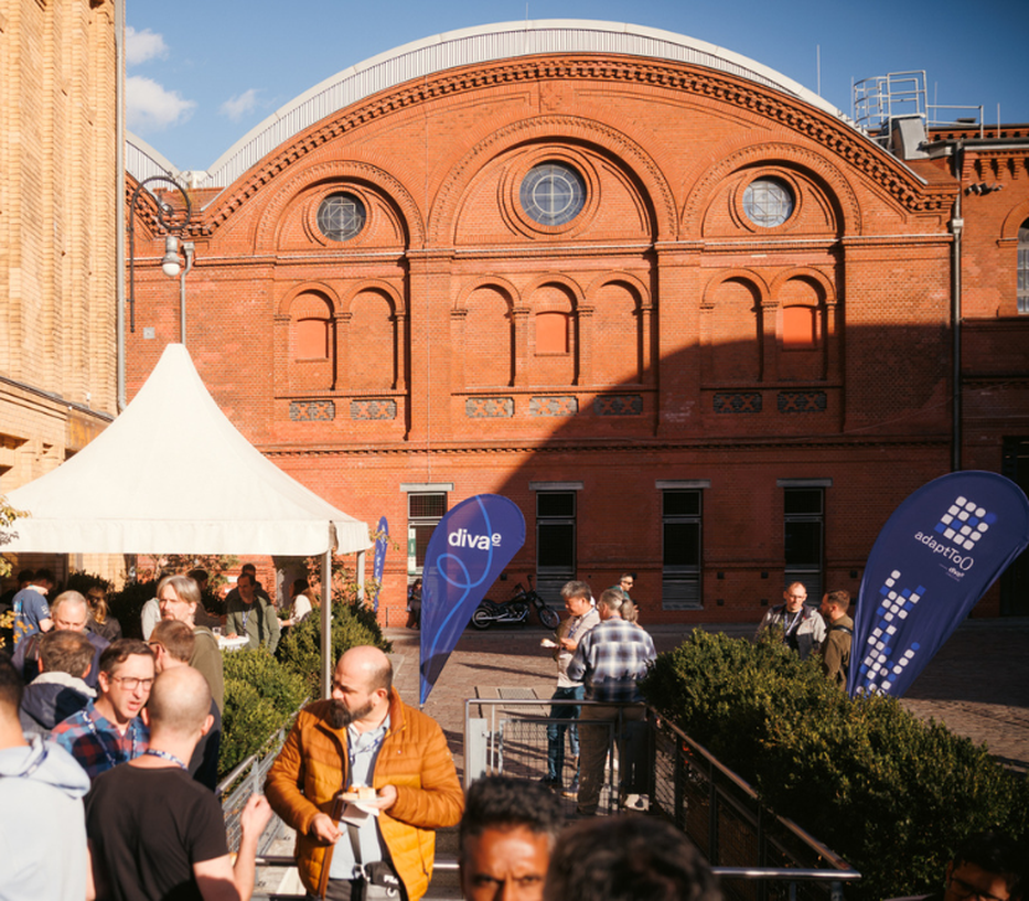 People gather under a tent in front of a historic red brick building with arched windows, surrounded by blue flags and greenery.