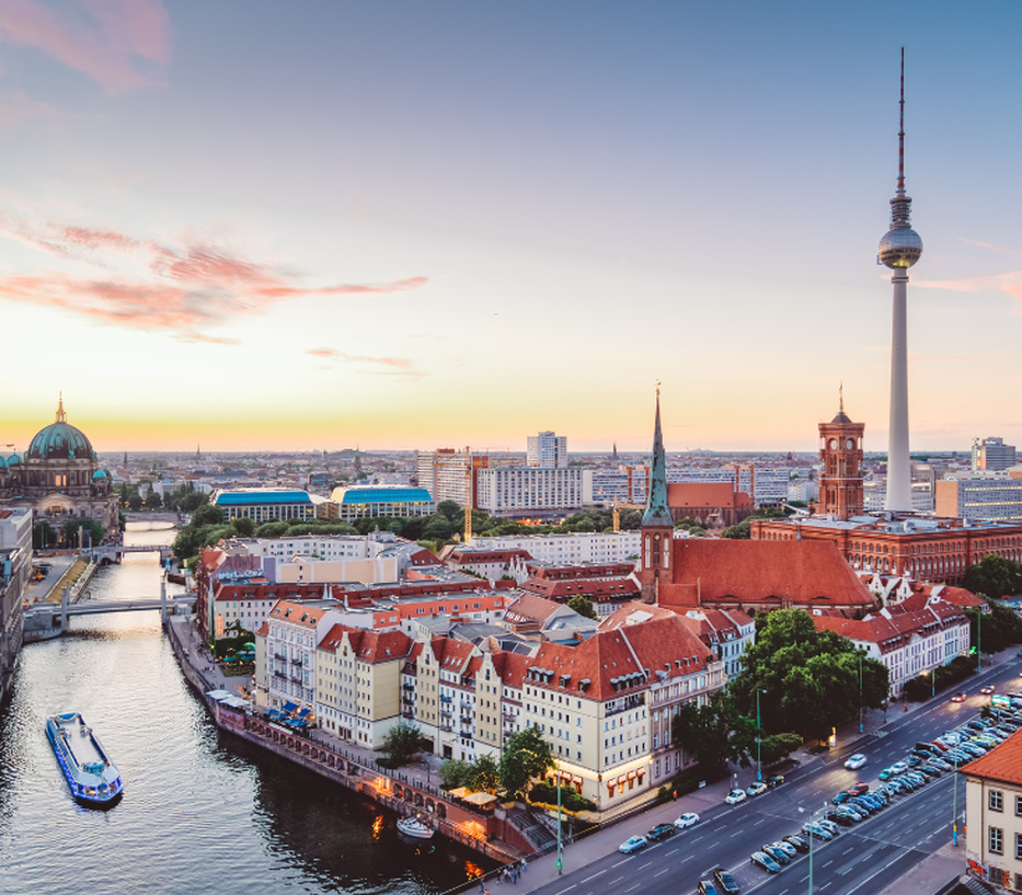 Aerial view of Berlin at sunset, featuring the TV Tower, river, historic buildings with red roofs, and a boat on the water.