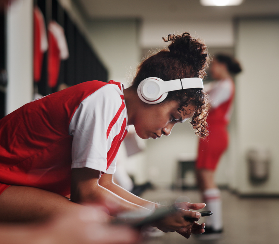 Soccer player in a red and white uniform sits in a locker room, wearing headphones and focused on a device. Another player is blurred in the background.