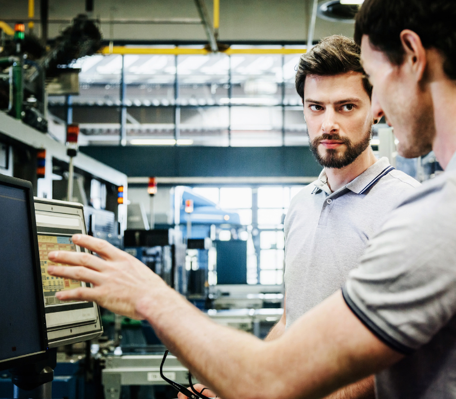 Two men in a factory setting, one pointing at a computer screen, discussing industrial machinery in the background.