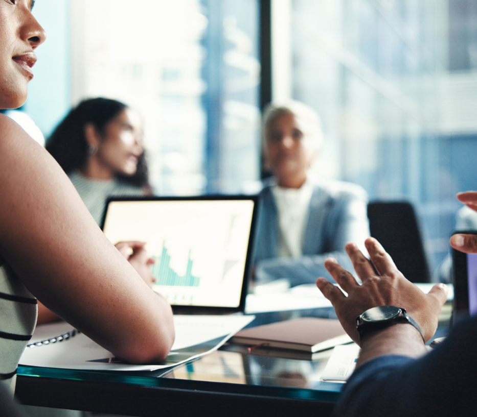 People in a meeting room discussing with a laptop displaying a graph on the table. Two blurred figures in the background.