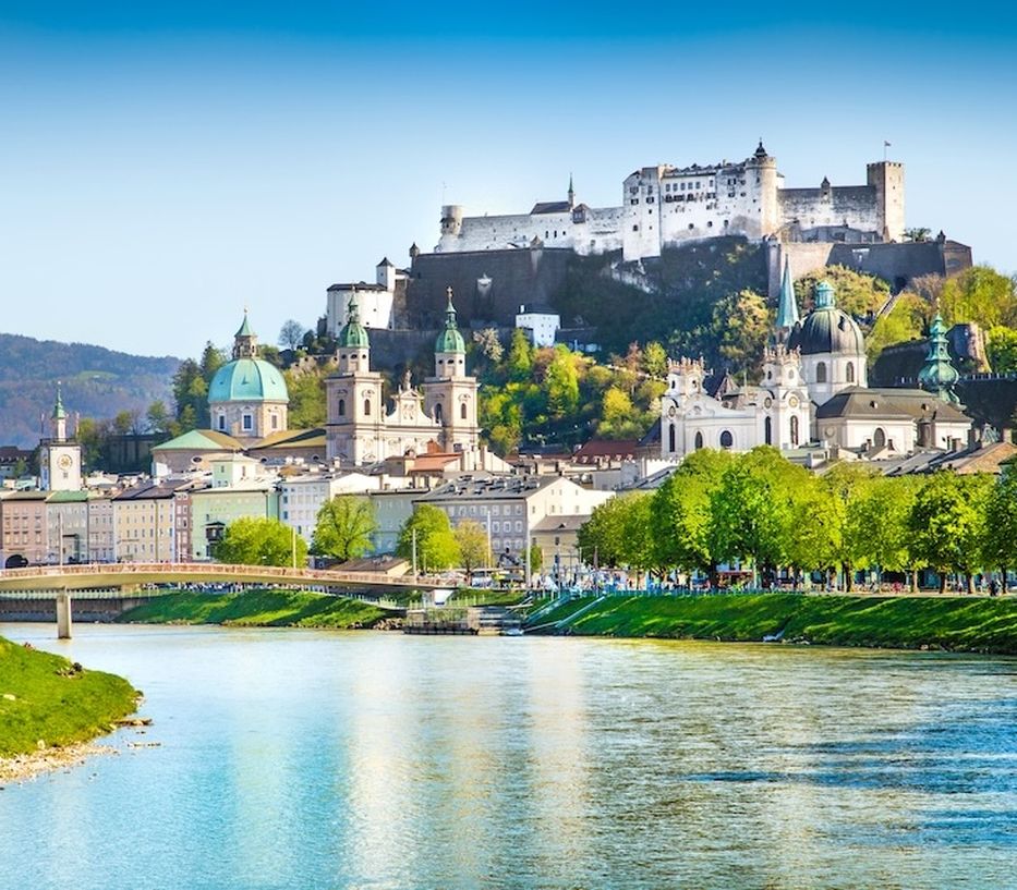 View of Salzburg, Austria, featuring the Hohensalzburg Fortress atop a hill, with historic buildings and a river in the foreground.