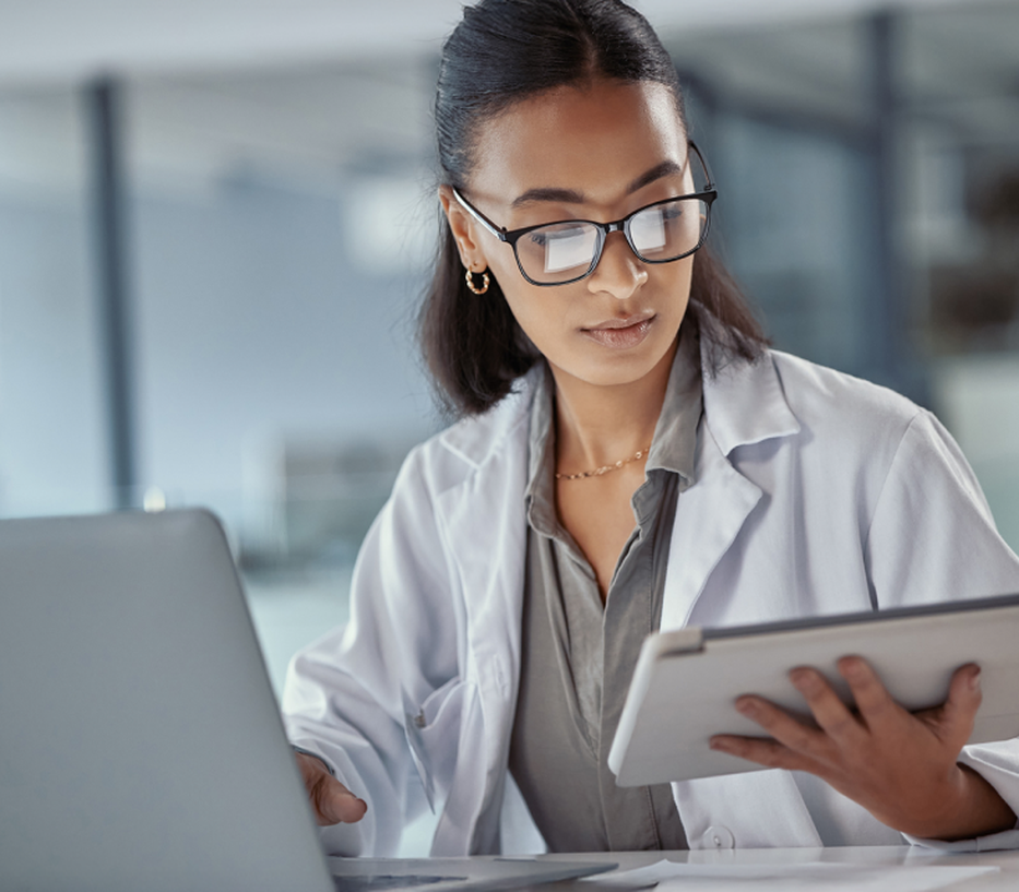 A woman in a lab coat and glasses works on a tablet and laptop in a modern office setting.