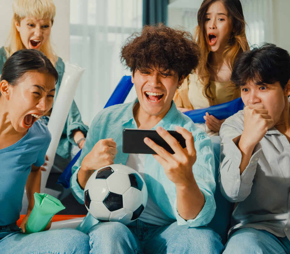 A group of excited friends watching a sports match on a smartphone, holding a soccer ball and cheering energetically in a living room.