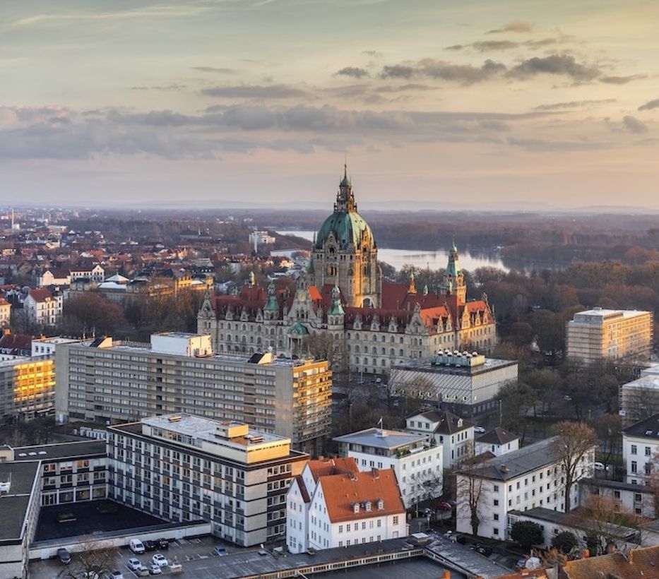 Aerial view of a city with a large historic building, intricate dome, surrounded by modern structures and tree-lined river in the distance at sunset.