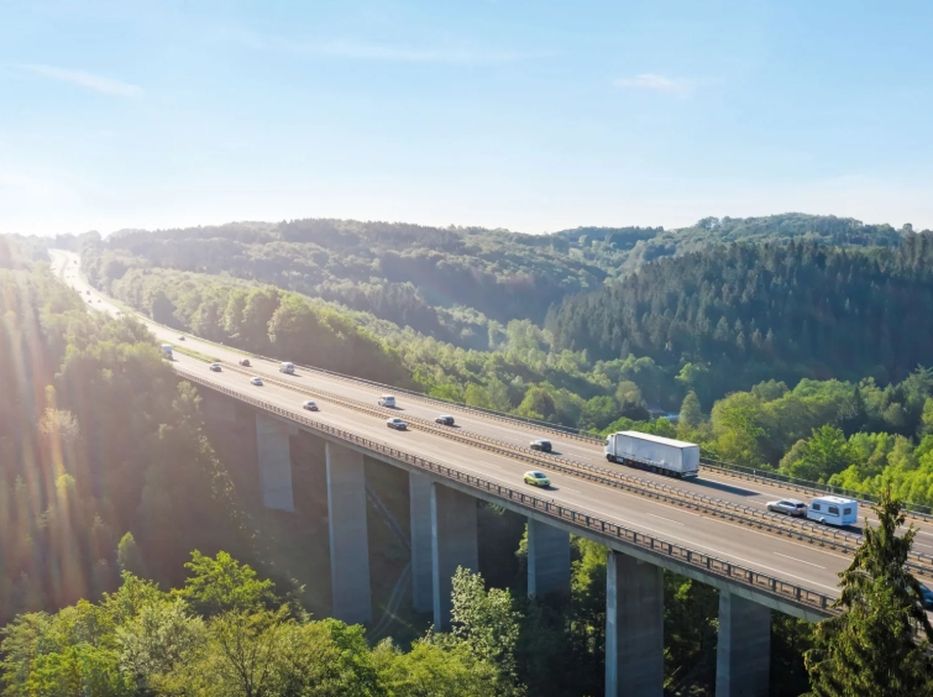 Aerial view of vehicles on a highway bridge surrounded by lush green forest and hills under a clear blue sky.