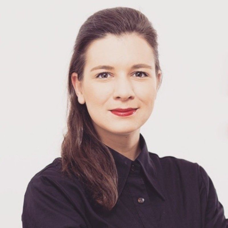 Woman with dark hair and red lipstick, wearing a black shirt, against a plain white background.