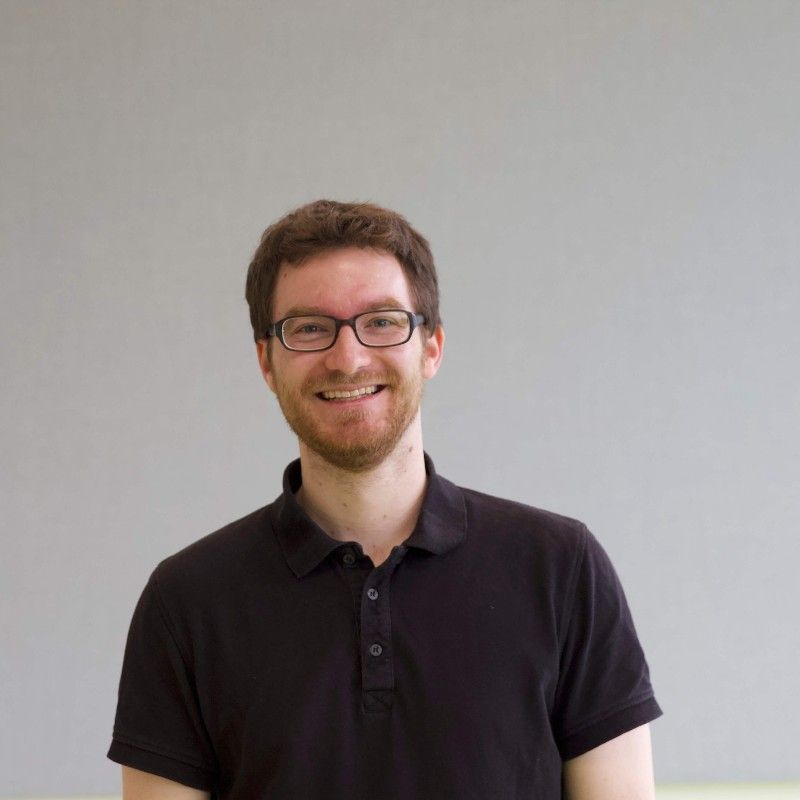 A smiling man with glasses and a beard, wearing a black polo shirt, stands against a plain gray background.