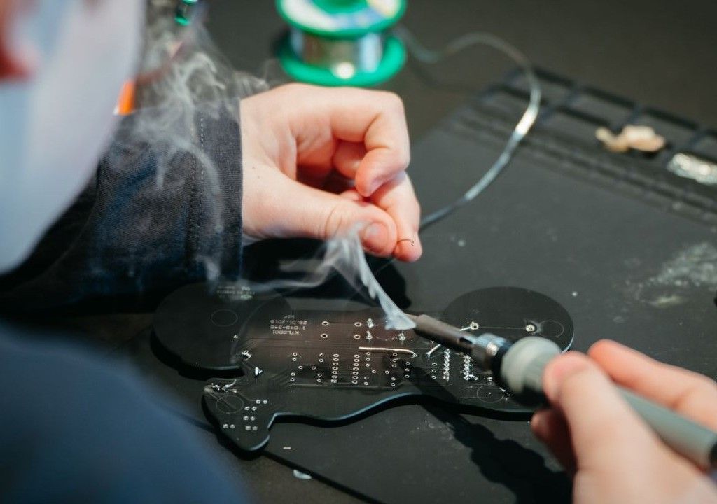 A person soldering components onto a black circuit board with a soldering iron, surrounded by smoke and electronic tools.