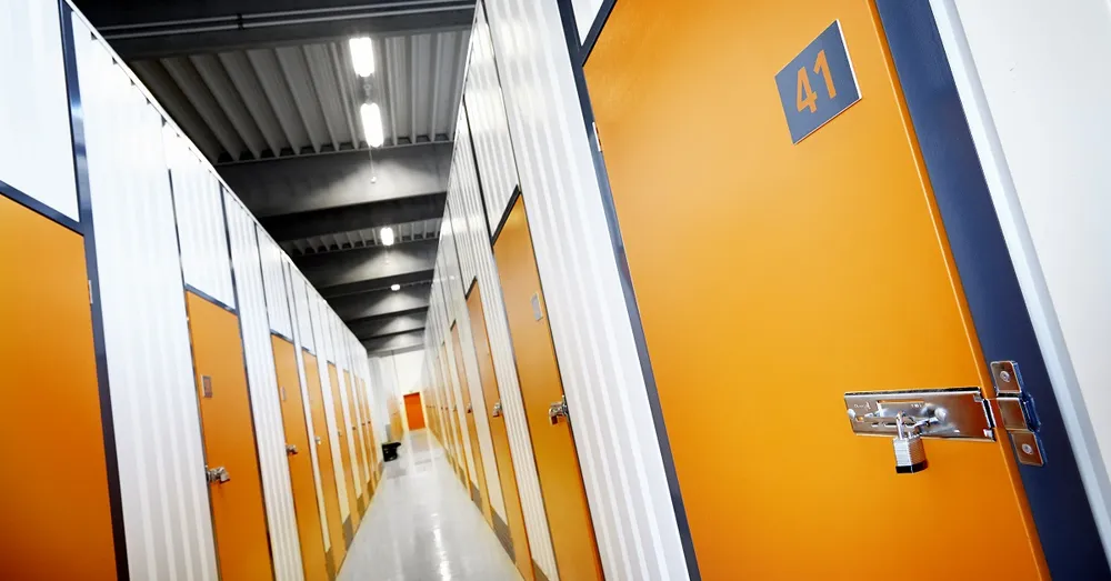 A corridor with rows of storage unit doors, painted orange and numbered, under fluorescent ceiling lights.