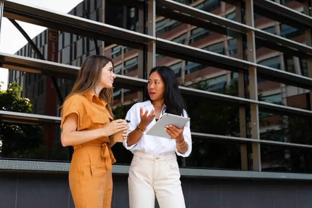 Two women are talking outside a modern building; one holds a cup, the other a tablet. They're dressed in casual, stylish clothing.