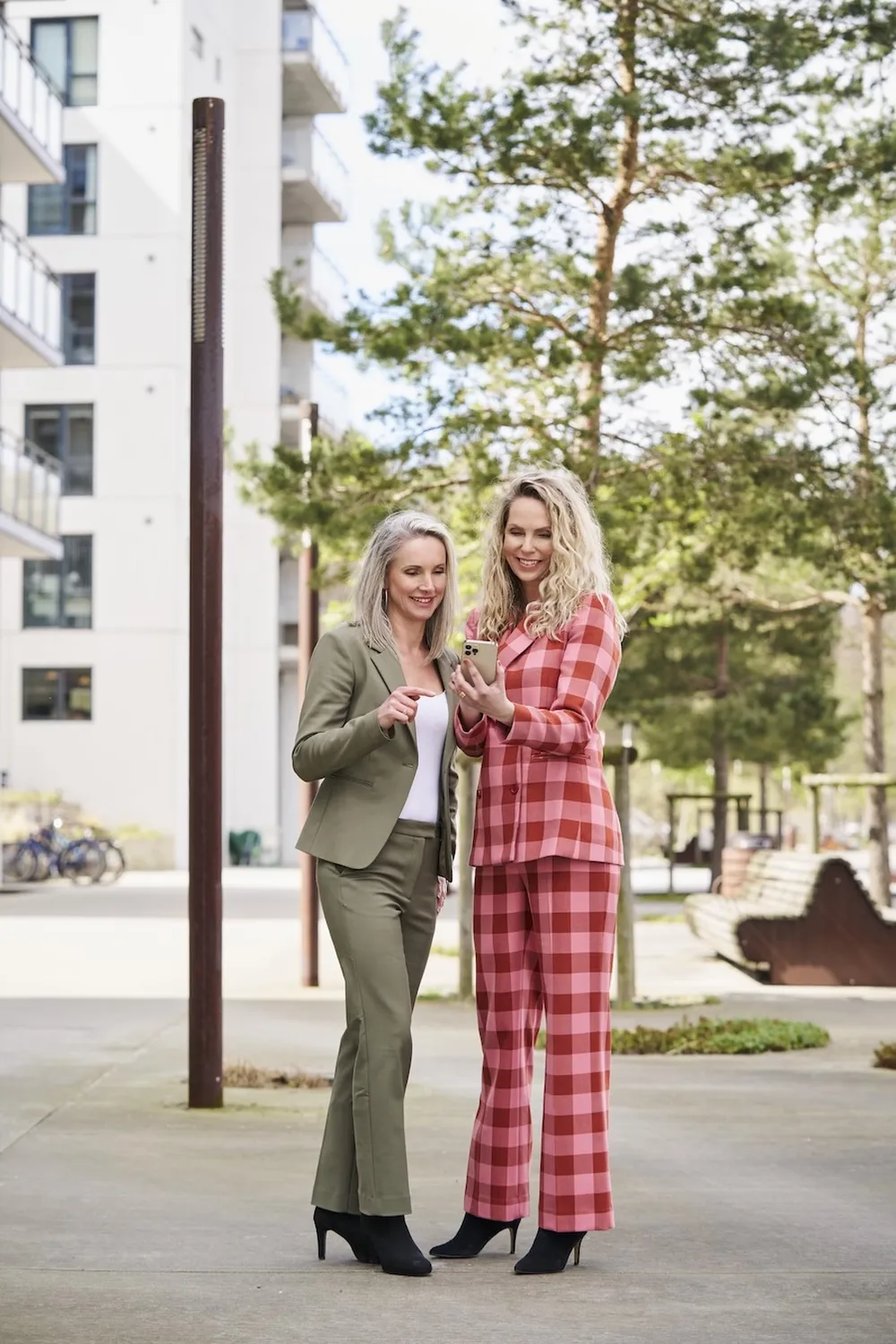Two women outdoors, one in a green suit and the other in a red plaid suit, smiling and looking at a smartphone. Trees and buildings in the background.