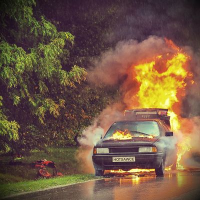 Cover of the Album "ENJOY THE FLAMES" depicting a Car with "HOTSAWCE" plate engulfed in flames on a rainy road, with trees in the background.