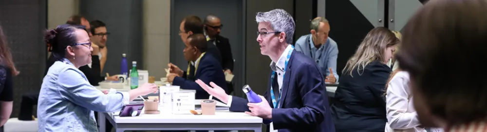 Two people in foreground talking across a table; others converse at nearby tables in a busy networking or conference setting.