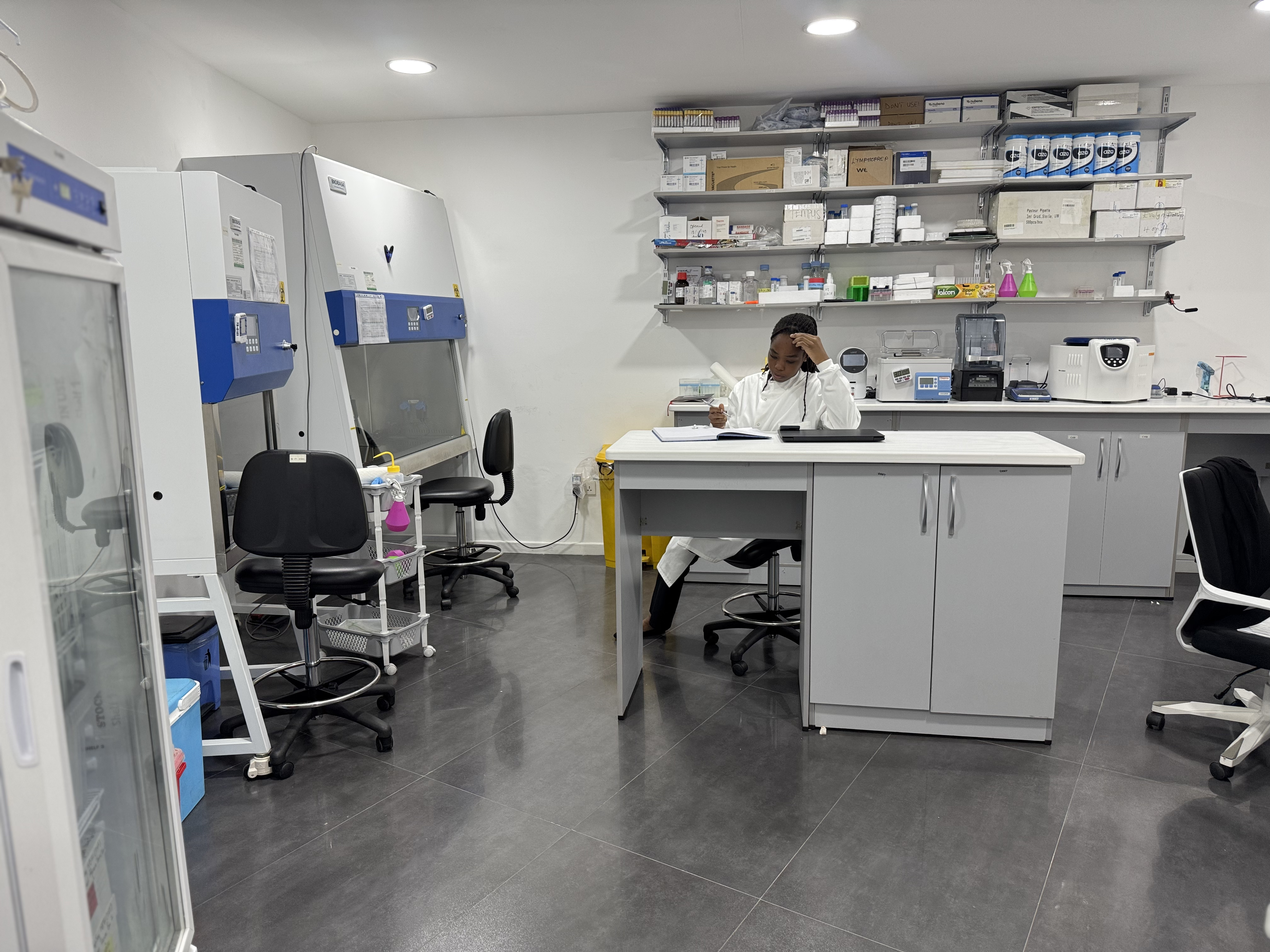 A woman in a lab coat is sitting on a white lab at a desk in the middle of a room, thinking whilst looking at a notebook