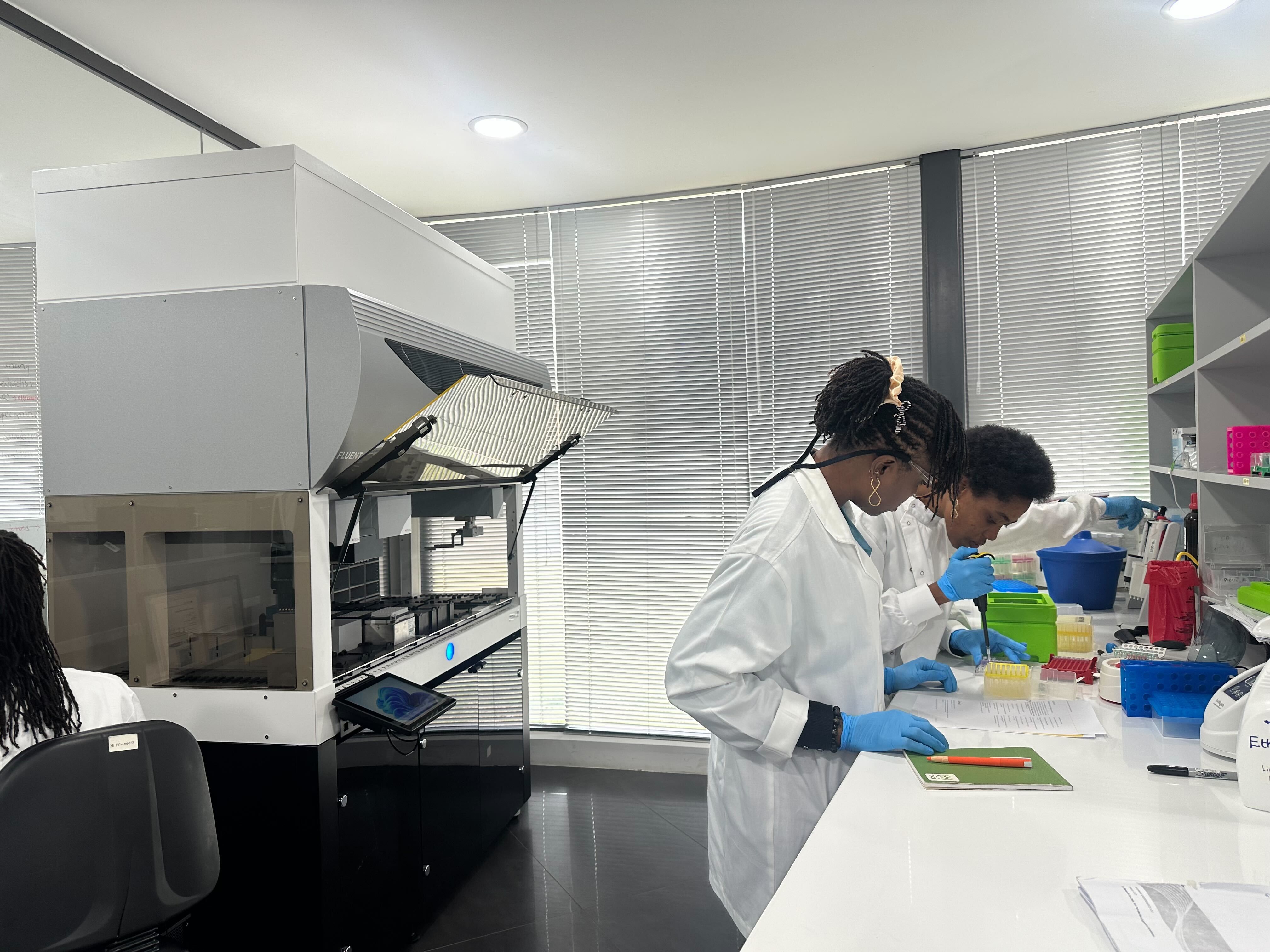 Two women are stood in a lab, one of them is pipetting and the other is watching with a notebook under her hand.