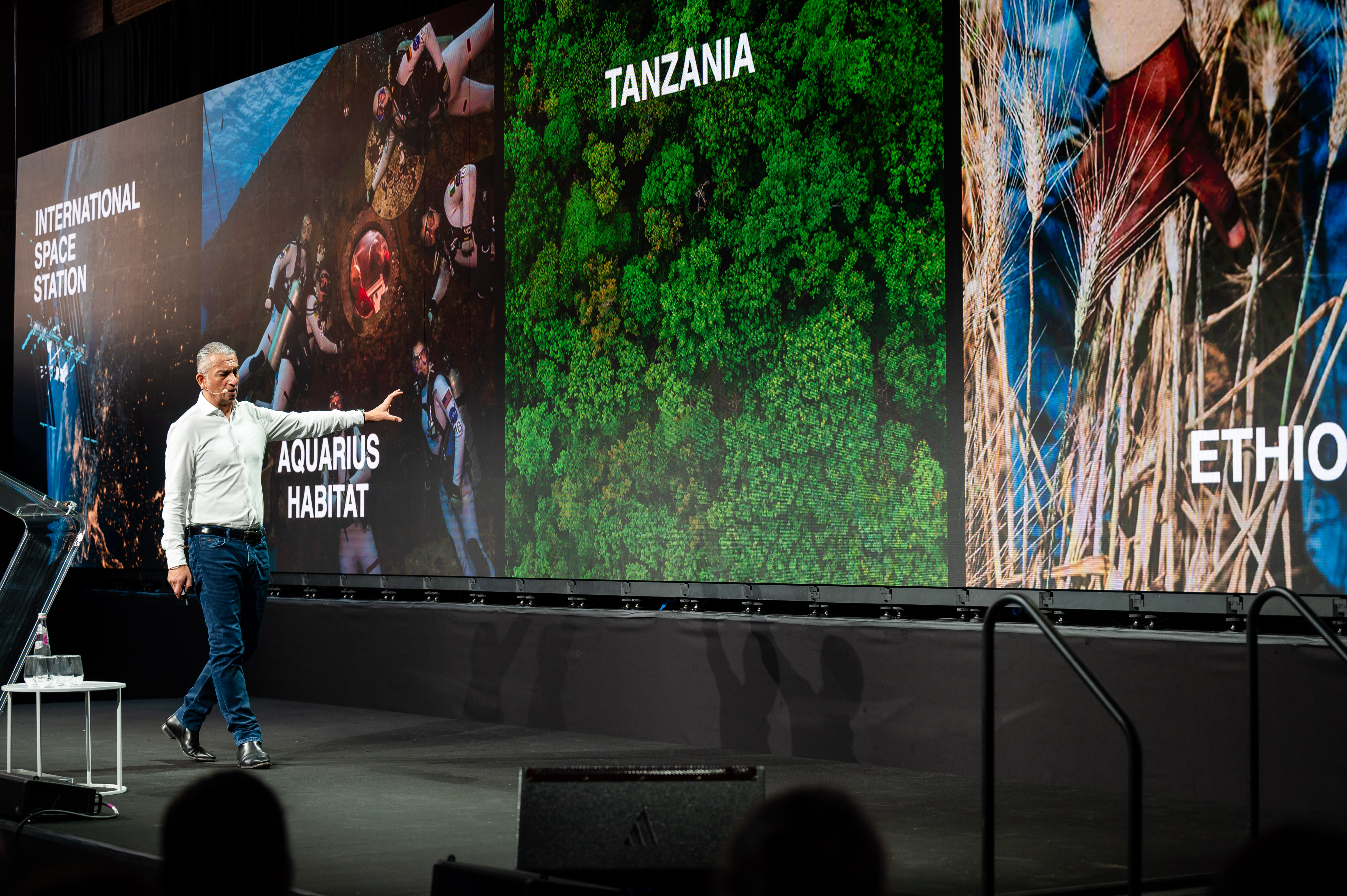 Gordon Sanghera walks across a stage in front of a screen showing a range of locations