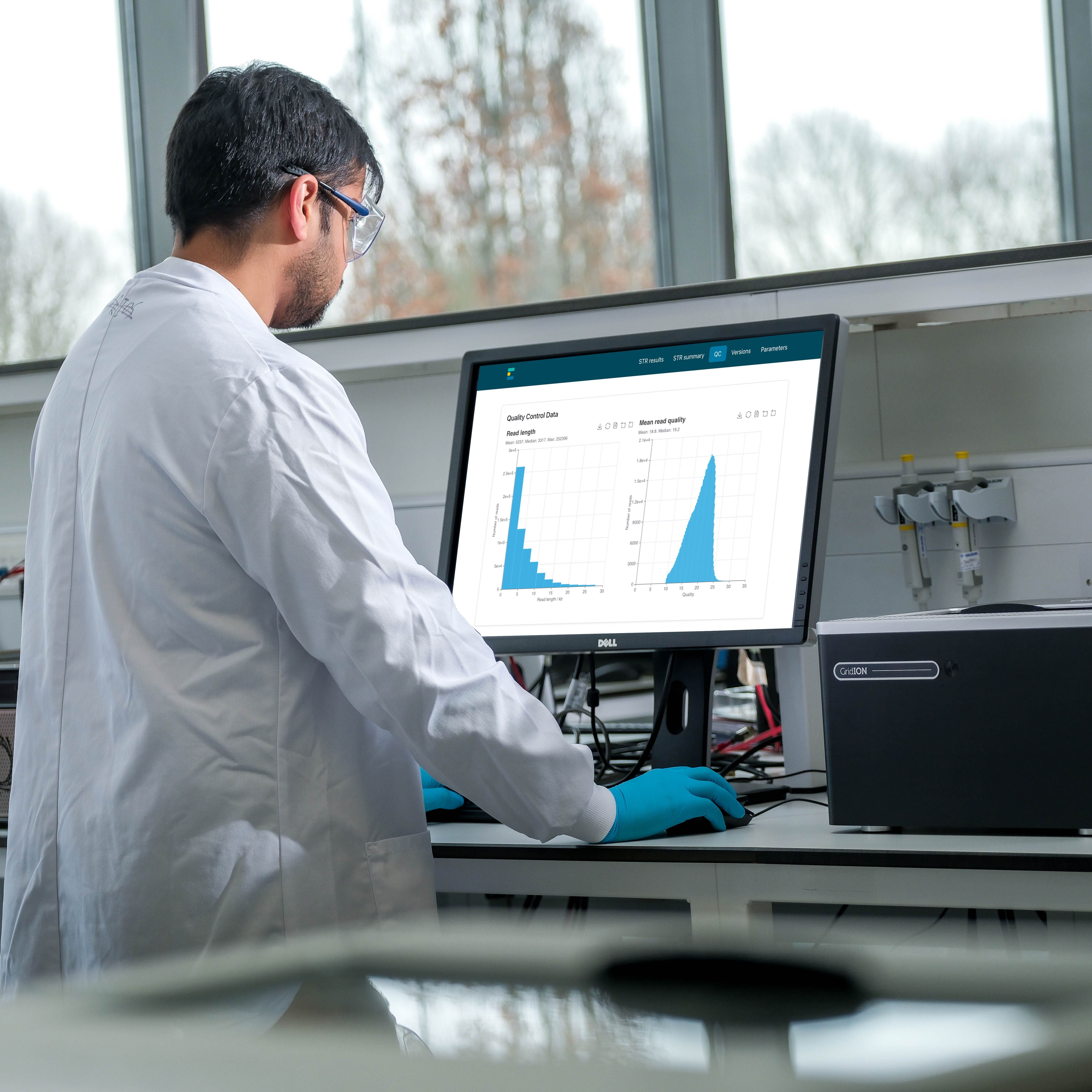 A Scientist wearing a white lab coat, goggles and gloves is on our computer looking at data within EPI2ME. They are in a lab and there are Oxford Nanopore sequencing devices to either side of them.