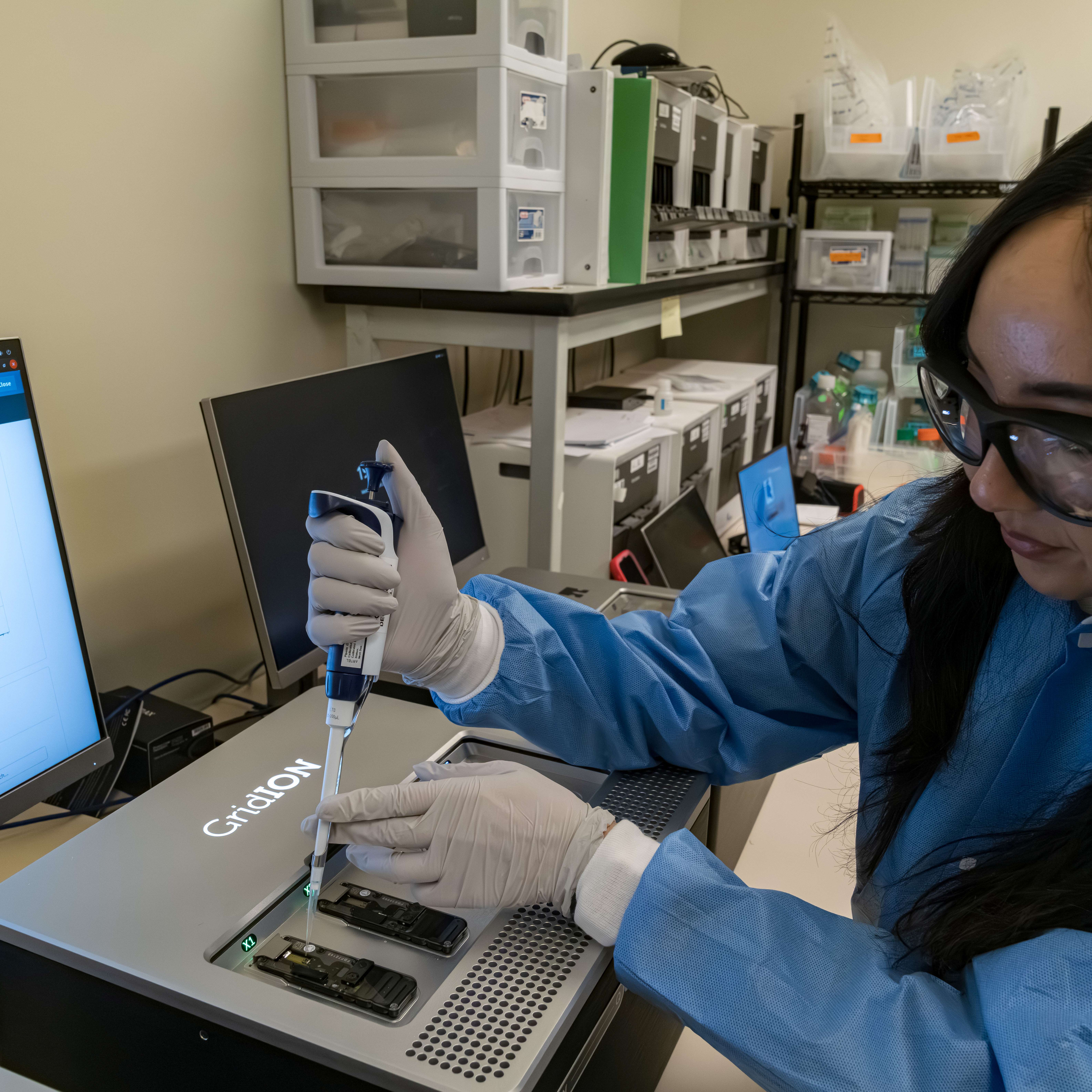Scientist in protective gear uses a pipette in a lab, analyzing results on a computer screen.