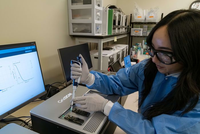 Scientist in protective gear uses a pipette in a lab, analyzing results on a computer screen.