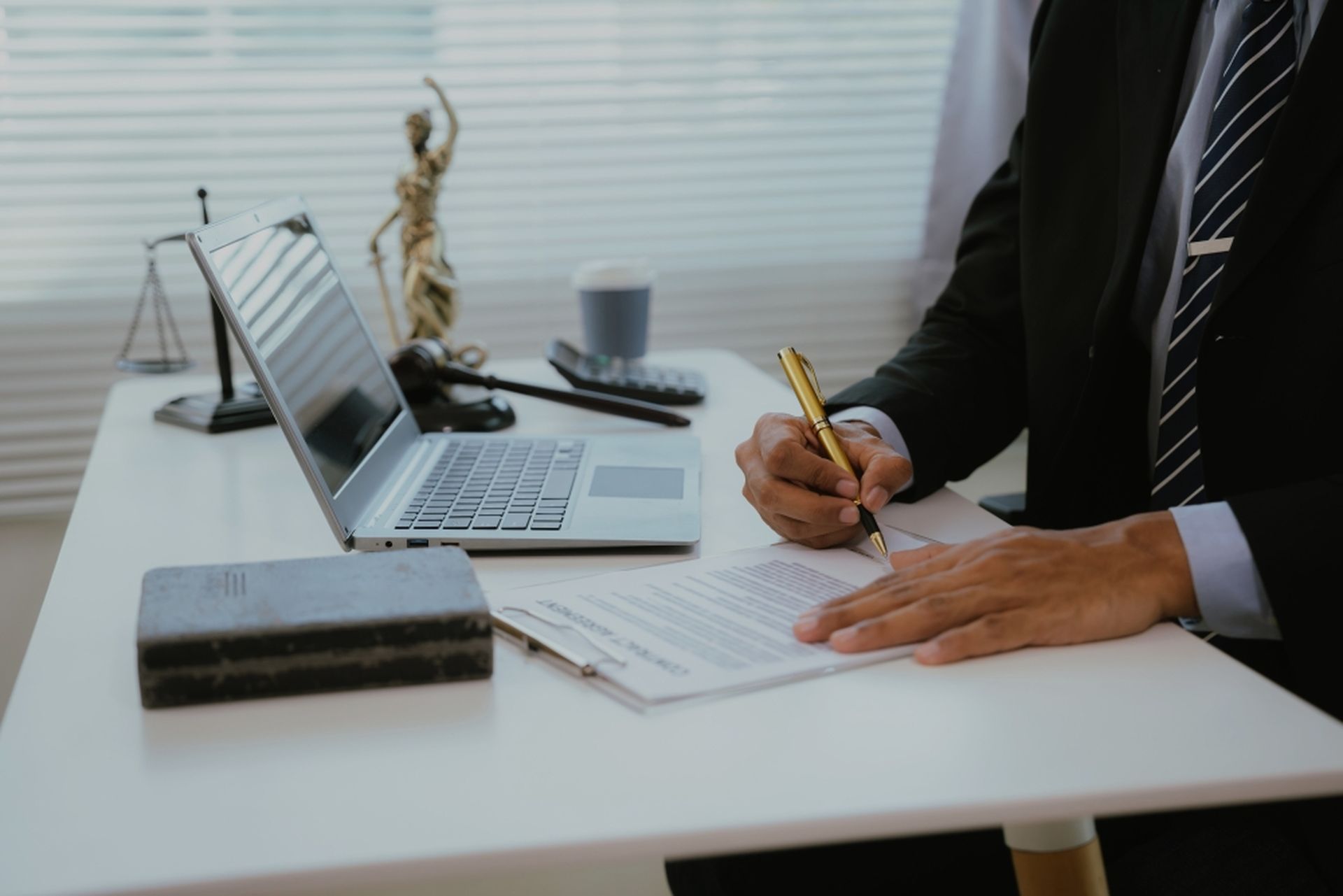 A person in a suit signing a document at a desk with a laptop, scale, statue, and book.