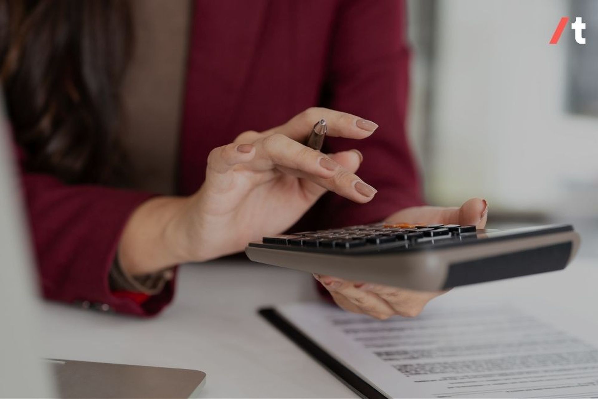 A person in a red blazer uses a calculator while holding a pen, with a document and laptop on the desk.