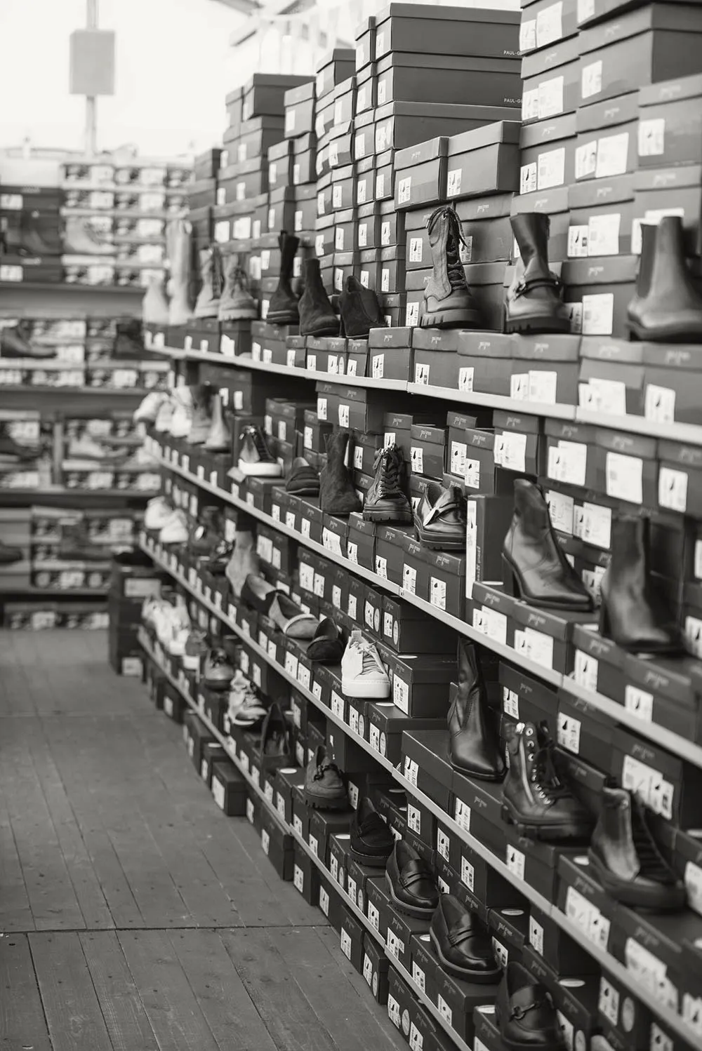 Reihen von gestapelten Schuhkartons, auf denen verschiedene Stiefel und Schuhe ausgestellt sind. Schwarz-Weiß-Aufnahme.