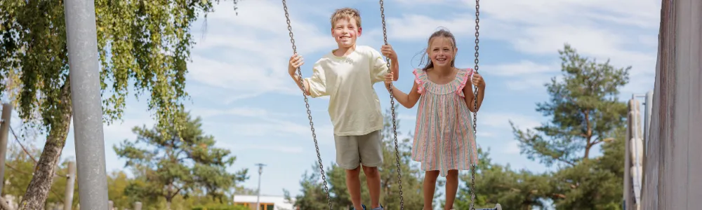 Zwei Kinder, ein Junge und ein Mädchen, die lächelnd auf einer Schaukel stehen, im Hintergrund ist ein sonniger Park zu sehen