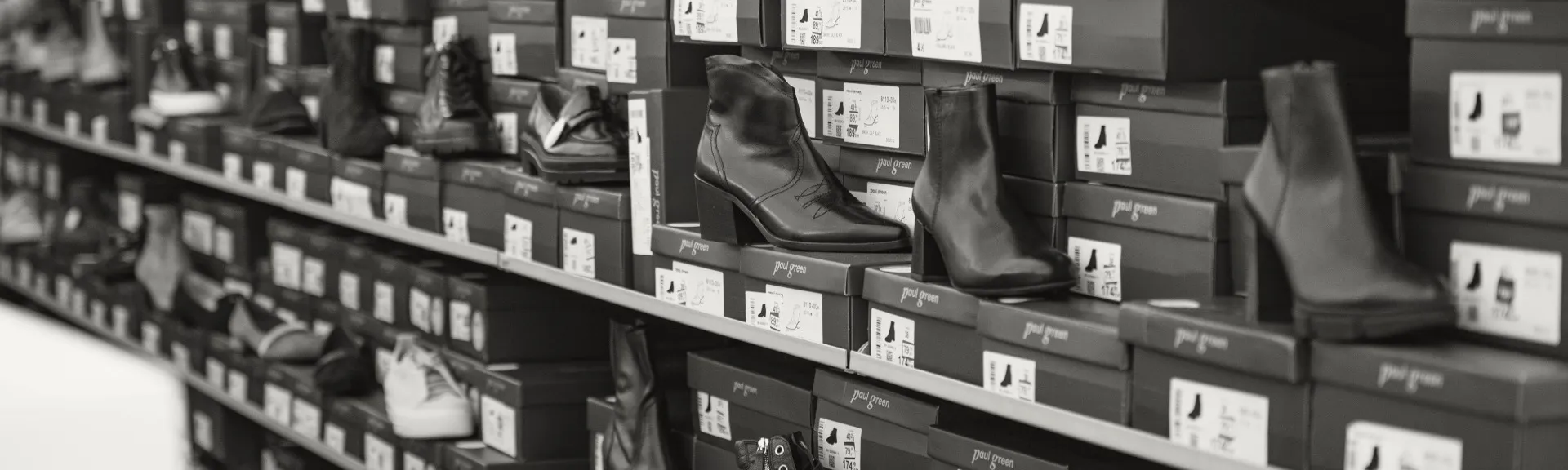 Ein Schwarz-Weiß-Foto von Regalen, die mit gestapelten Schuhkartons gefüllt sind, auf denen teilweise Stiefel mit hohen Absätzen ausgestellt sind.