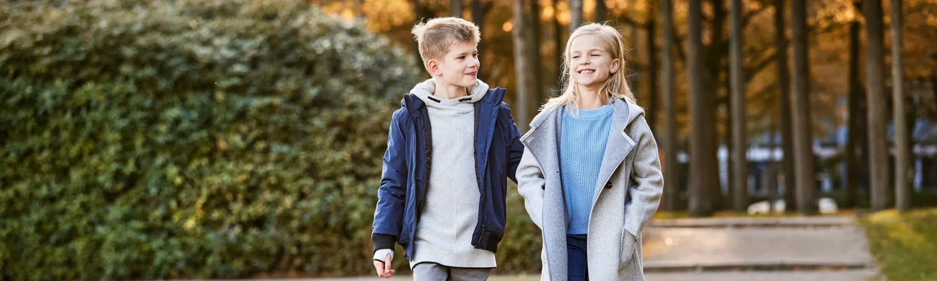Zwei Kinder gehen gemeinsam auf einem Weg durch einen herbstlichen Wald, beide tragen Jacken und lächeln einander an.