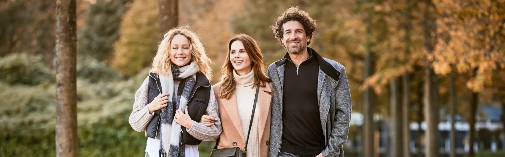 Three people smiling and walking together outdoors in a park during autumn, with trees and foliage in the background.