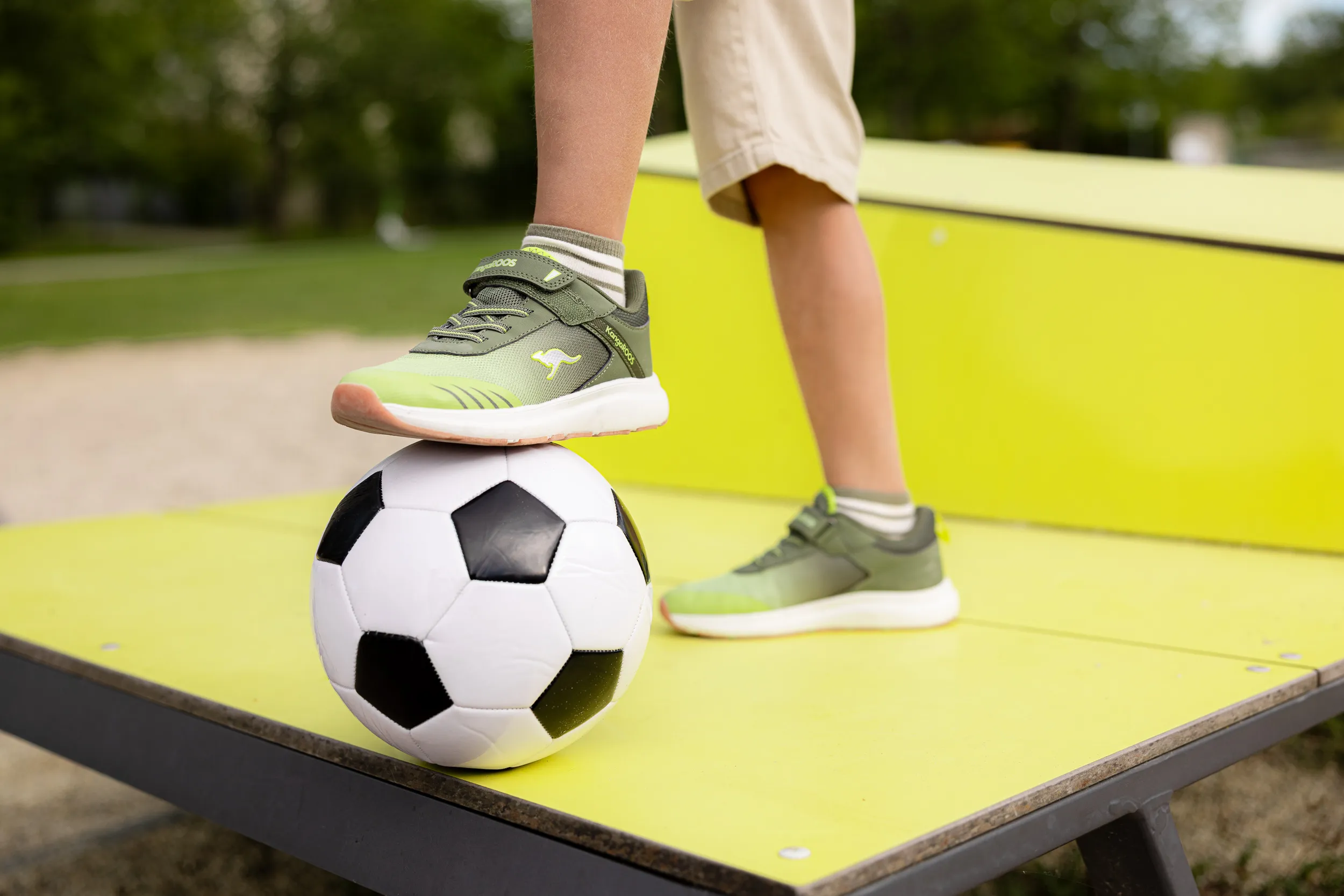 Kinderfüße mit grünen KangaROOS-Sneakern auf einem Fußball auf einem Spielplatz.
