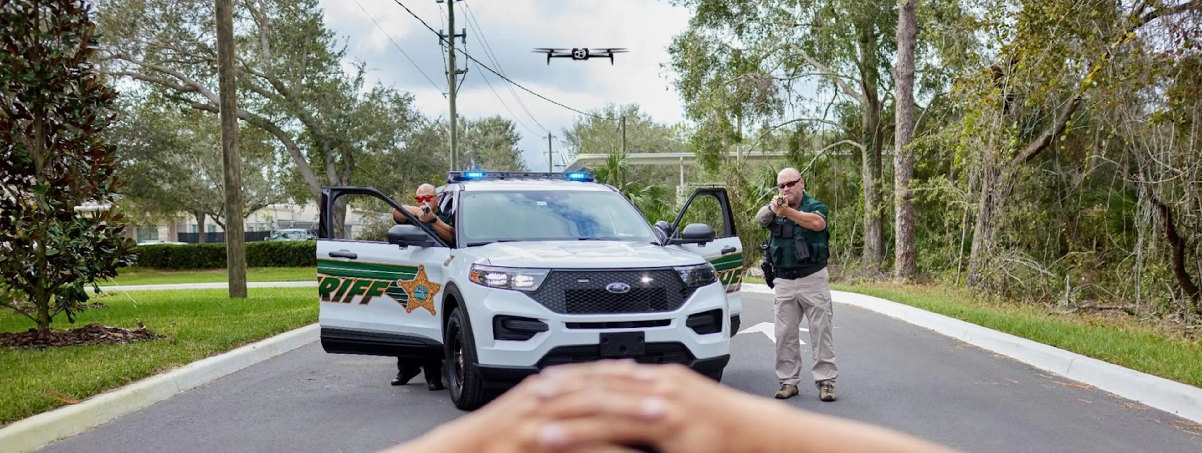 Law enforcement officers detain a suspect while a drone hovers over their vehicle.