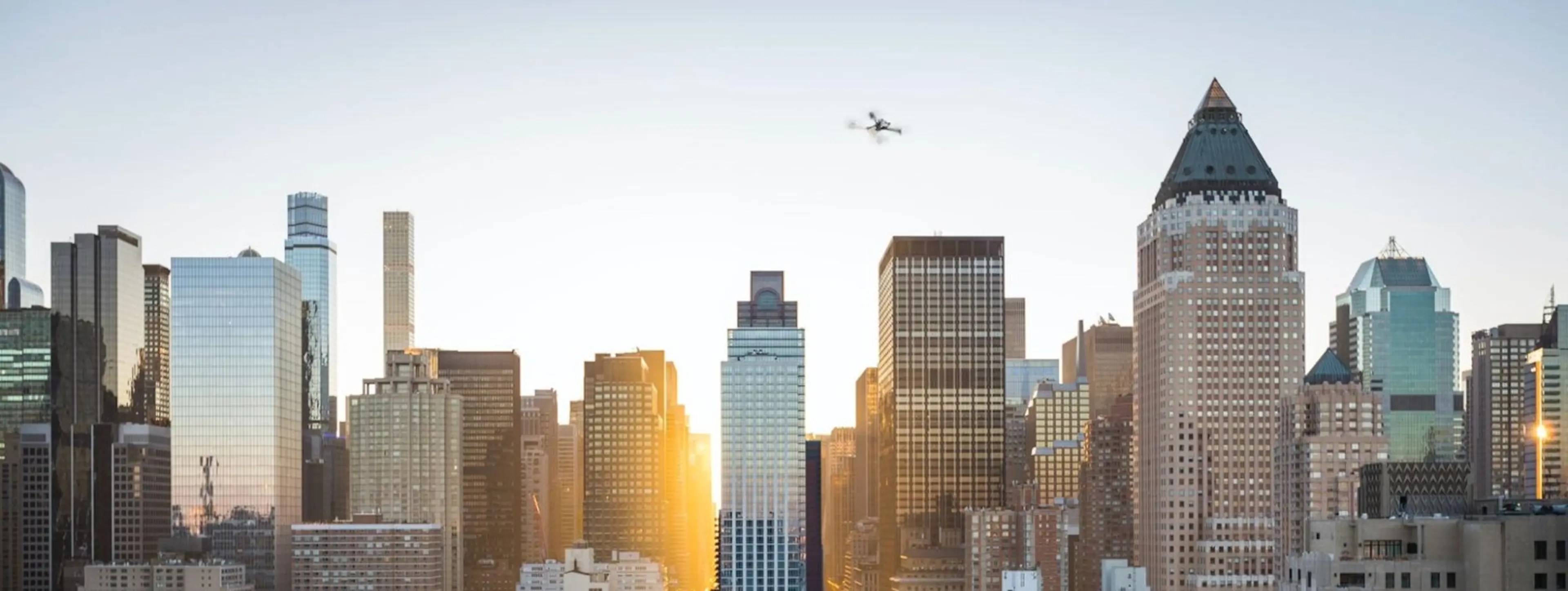 A drone flies high above a bustling cityscape as sunlight peeks around skyscrapers.