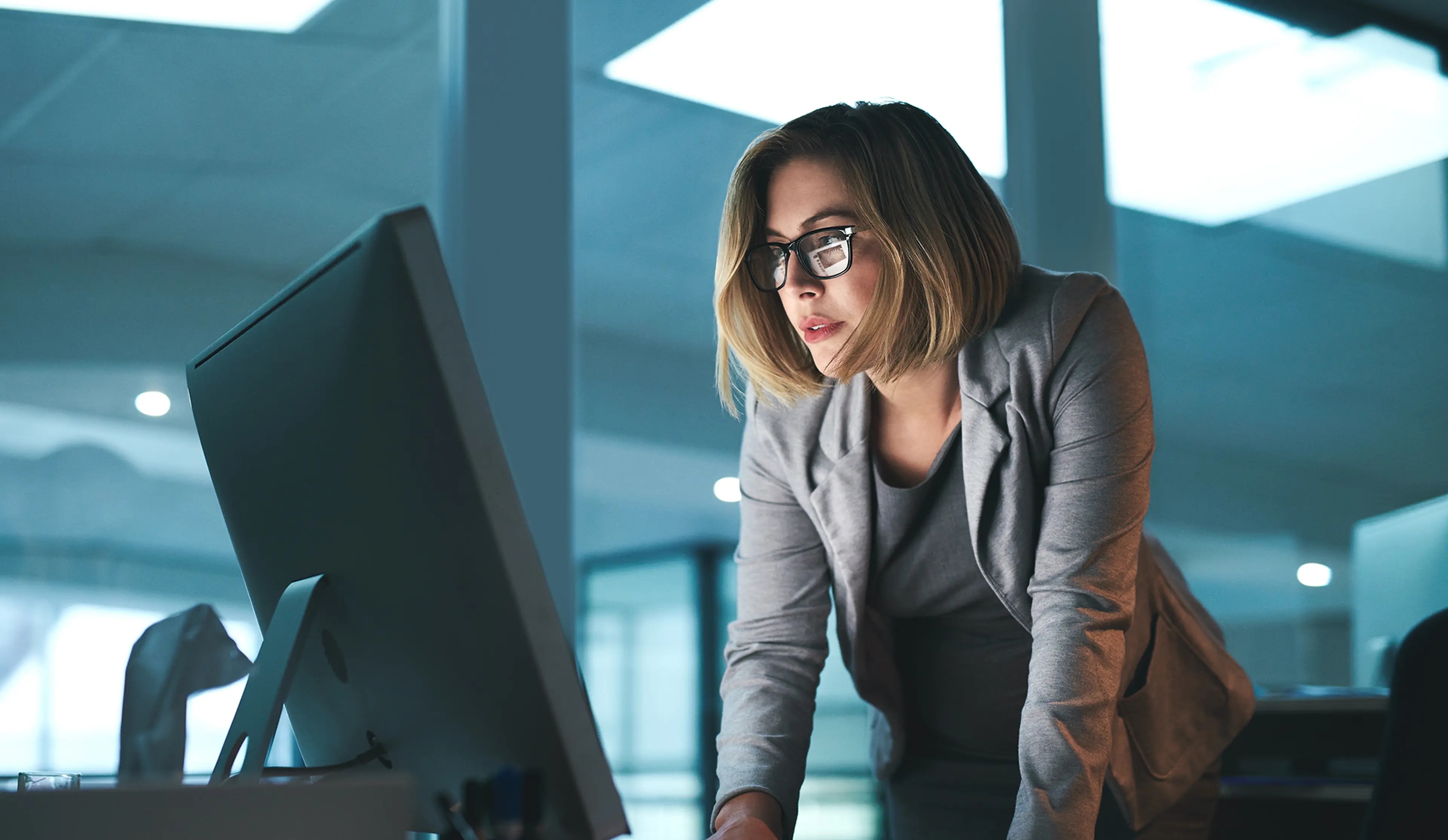 A woman in a gray blazer and glasses leans over a desk, intently looking at a computer monitor in a modern office setting.