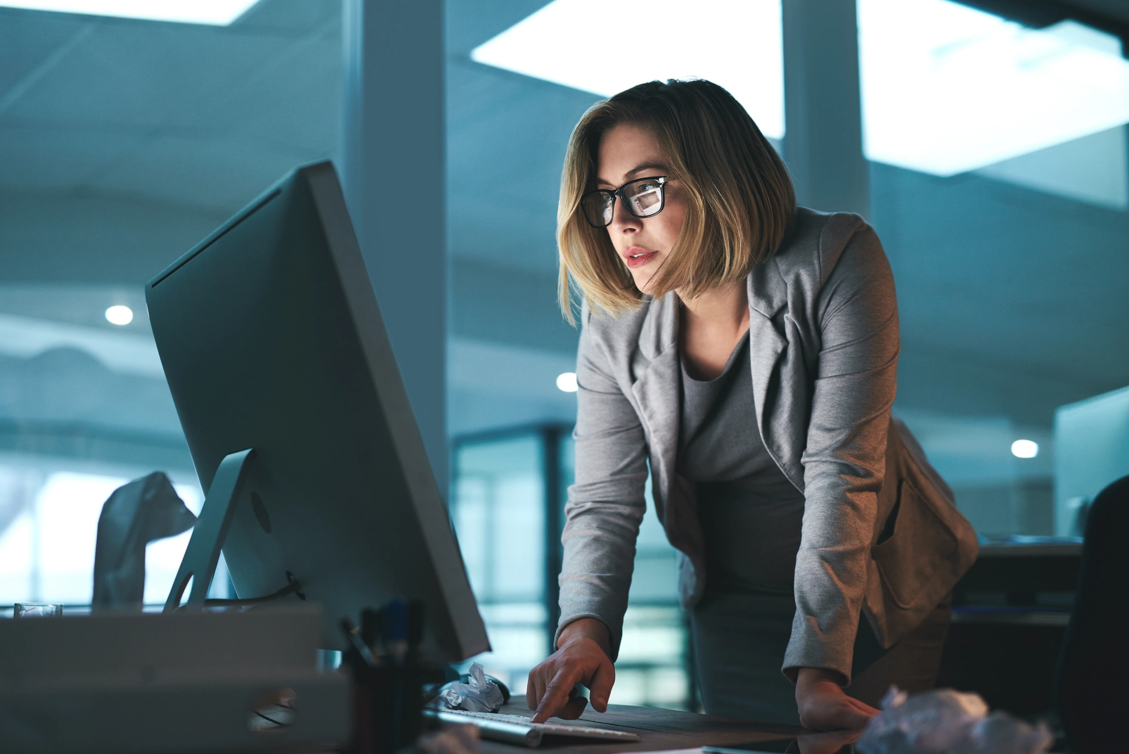 Woman in a gray blazer and glasses works intently on a desktop computer in an office with dim lighting.