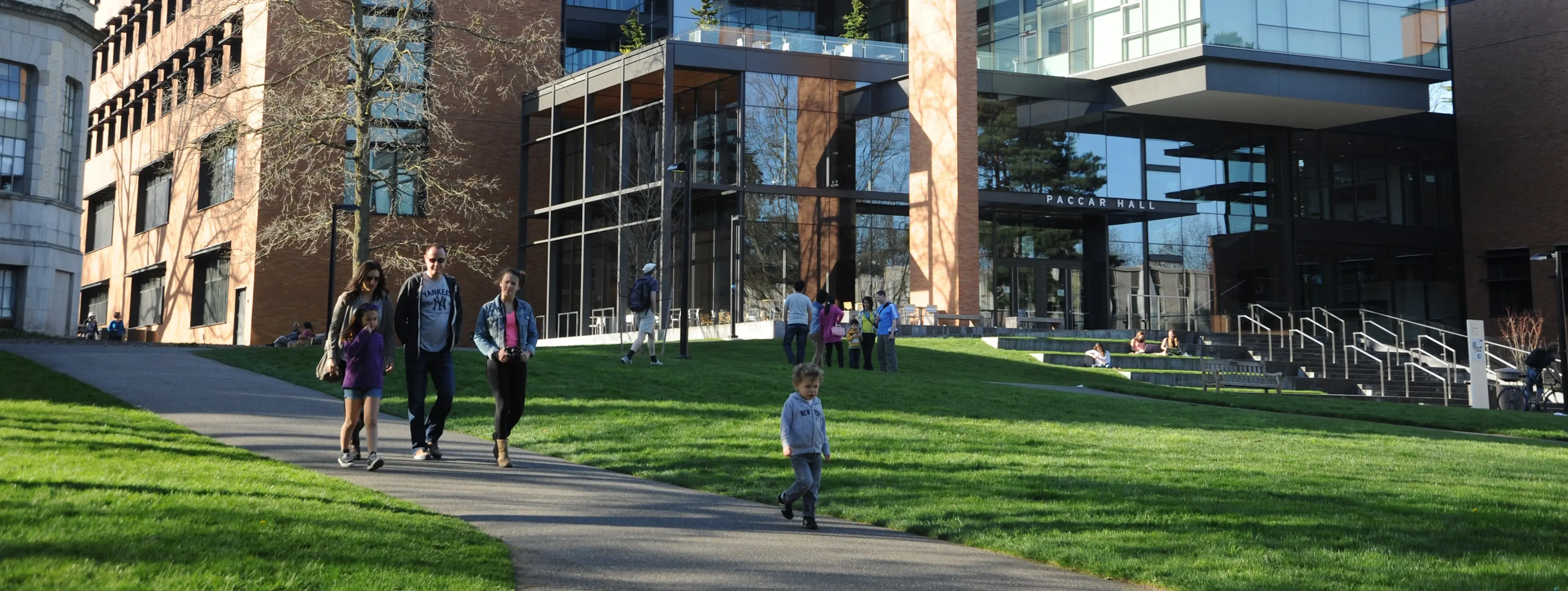 Exterior of a university building, with an expansive lawn. There is a group of students by the entrance and a family on a campus tour in the foreground.