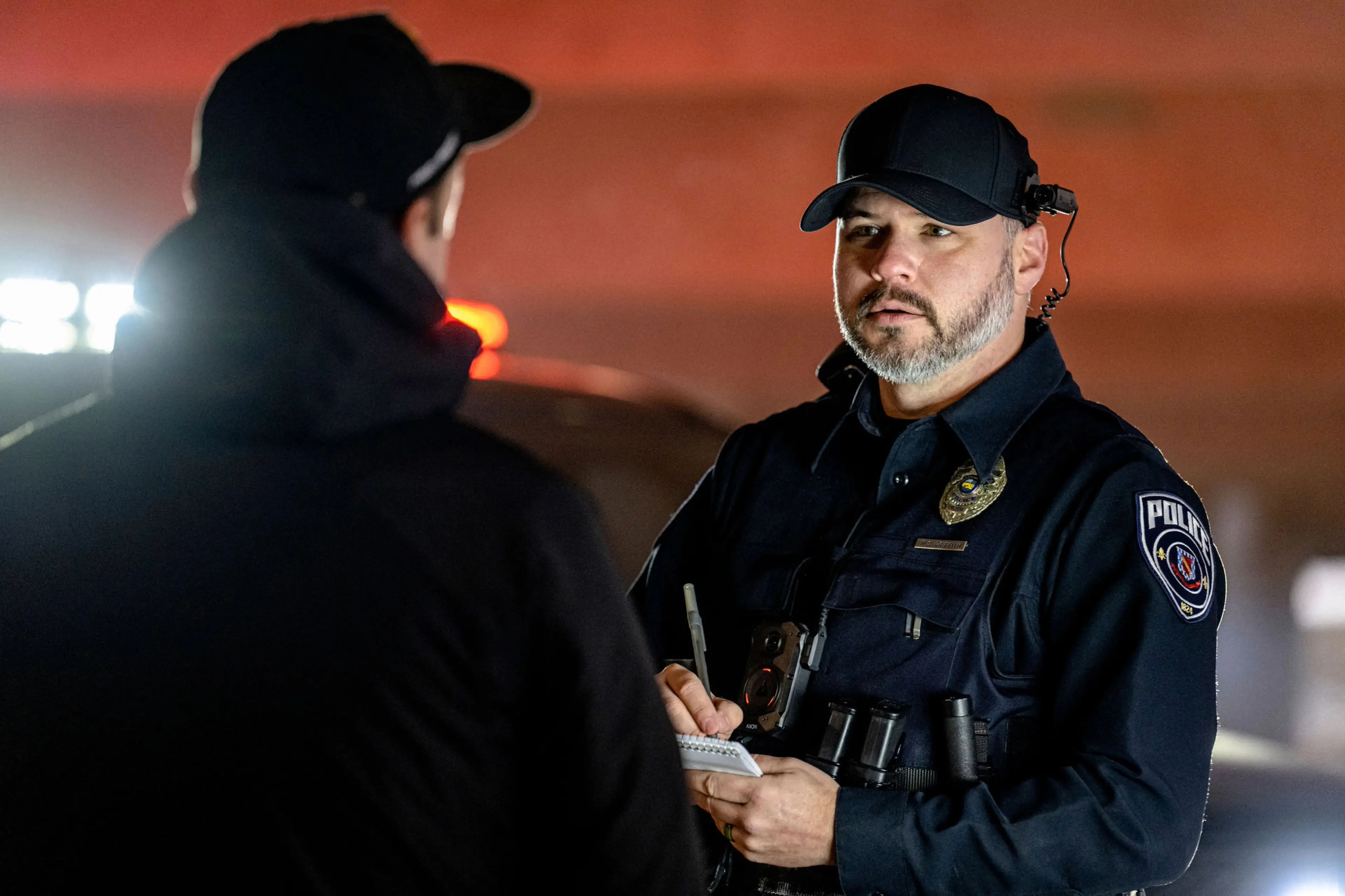 A police officer wearing an Axon Body 4 camera with POV attachment takes a witness statement at sunset.