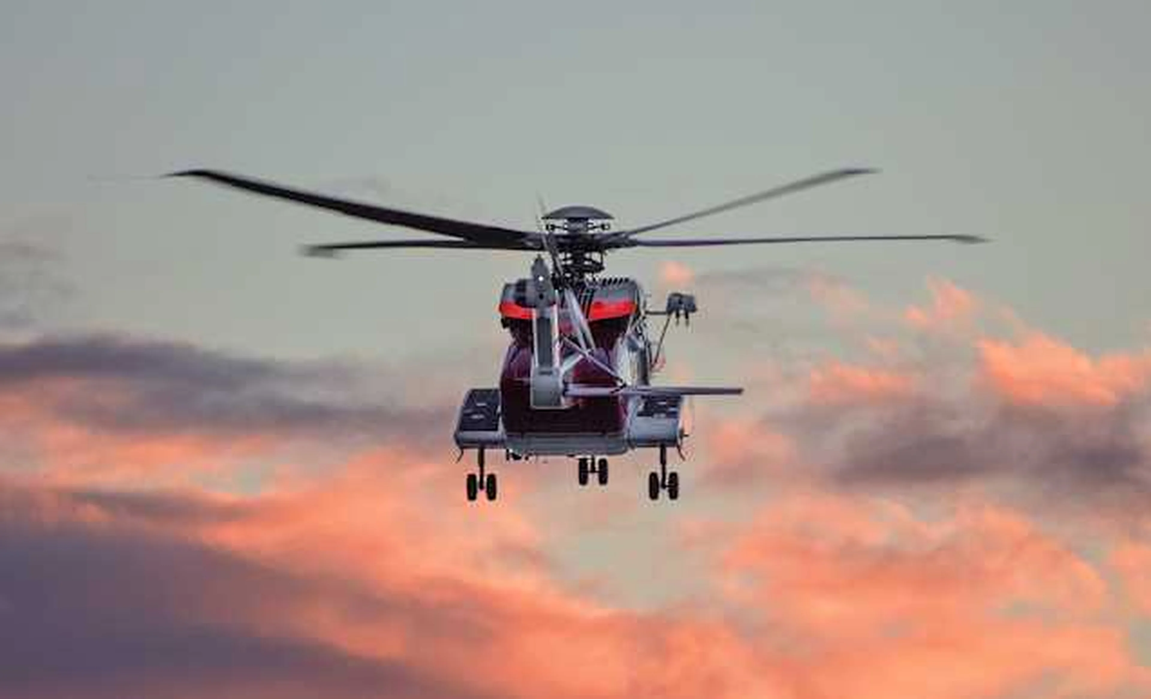 Rear view of an airborne, red-and-white rescue helicopter with a blue, pink, and grey sky in the background.