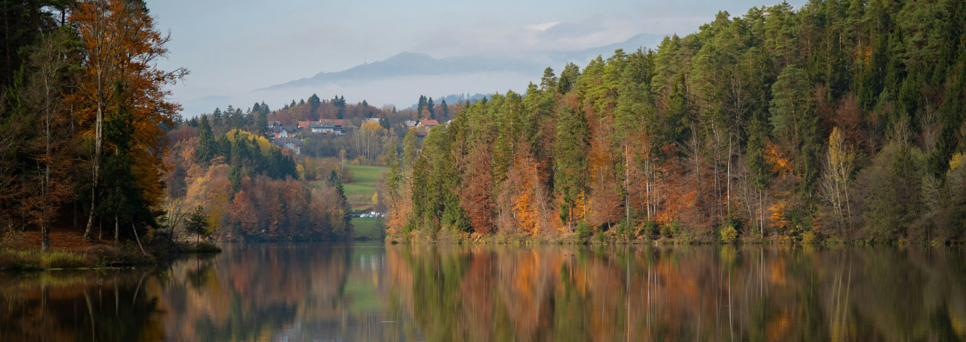 Serene lake reflecting autumn trees, a small village on distant hills and misty mountains in the background.
